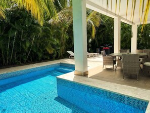 Une piscine dont les bords sont recouverts de carreaux, entourée d’une végétation luxuriante et dotée d’un espace extérieur aménagé, abrité et équipé de mobilier en osier.