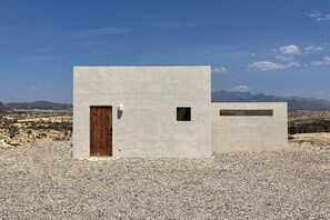 A modern building with a wooden door and a small window, set against a desert landscape with mountains in the background. - Adobe + Casita | Sleeps 6 | 10 Min to Big Bend (Terlingua)