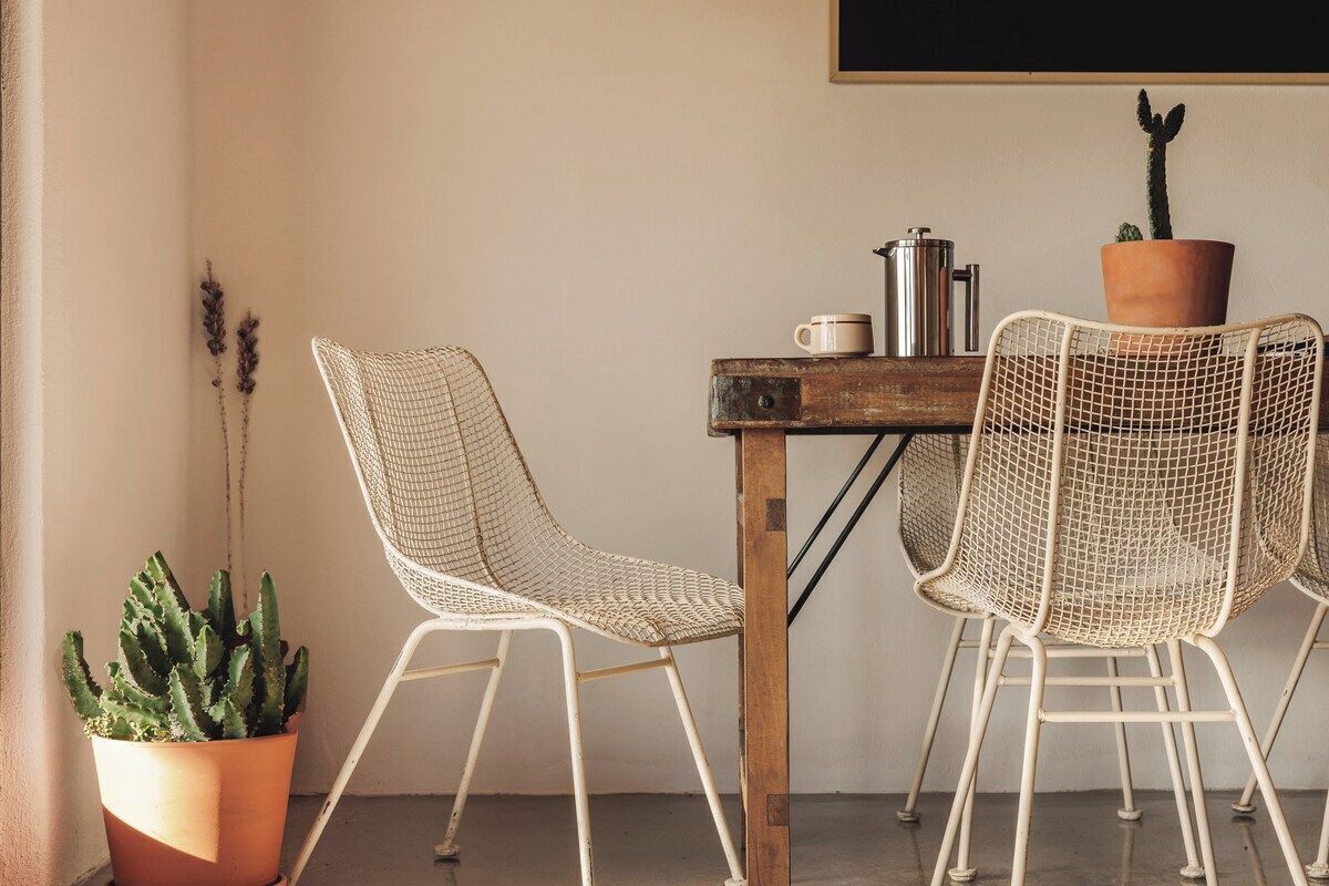 Une pièce comprenant une table et deux chaises en treillis métallique, un cactus en pot et une cafetière en métal.