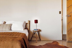 A bedroom with a bed, a wooden side table with books, a red lamp, and a wooden door. - Adobe + Casita | Sleeps 6 | 10 Min to Big Bend (Terlingua)