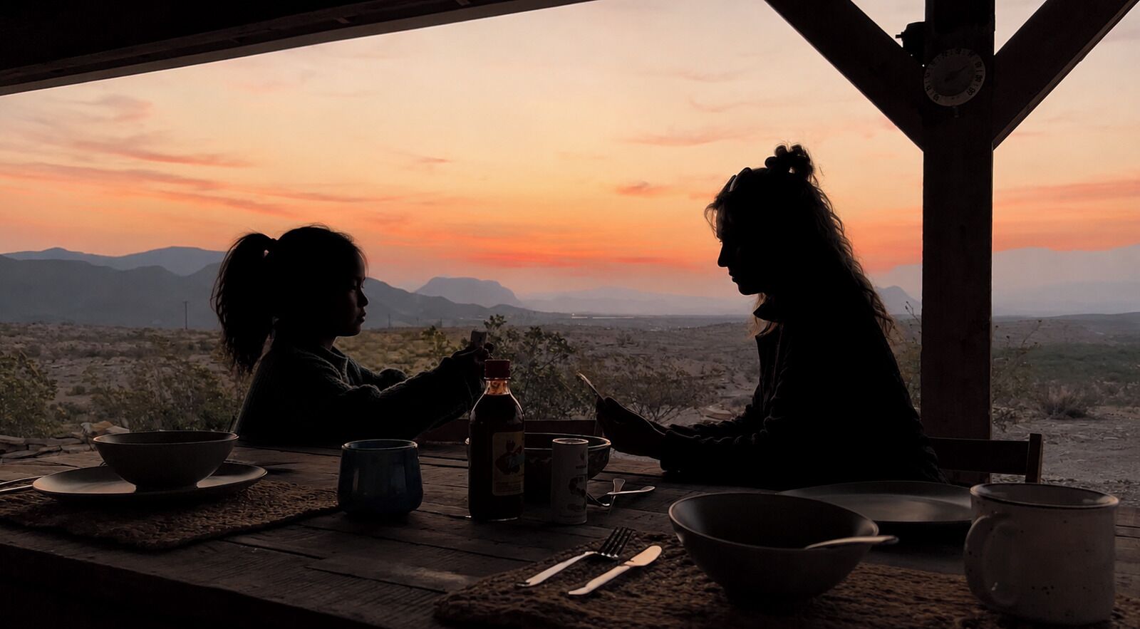 Deux personnes sont assises à une table en bois, devant laquelle s’étend un paysage de montagnes baignées par les couleurs du coucher de soleil.