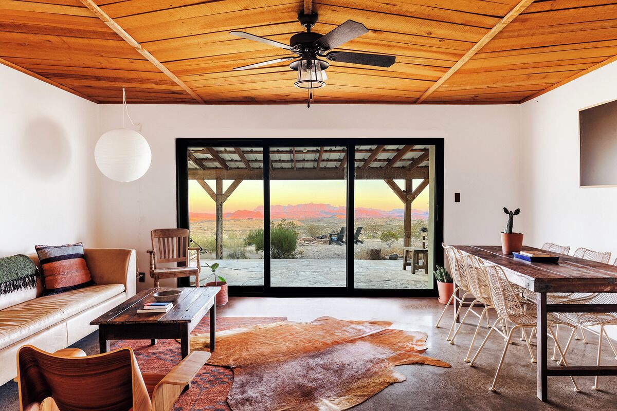 A spacious room with a wooden ceiling, a ceiling fan, a large sliding glass door leading to an outdoor view, a dining table with chairs, and a leather rug.