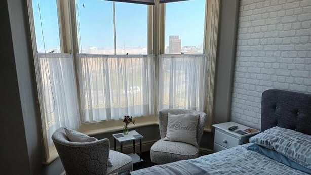 Una habitación de hotel con un ventanal grande, una cama, una silla, una mesita con un jarrón y una mesita de noche.