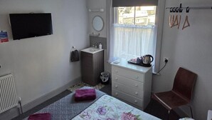 Un dormitorio con cama, ventana con cortina, cómoda, silla y televisor montado en la pared.