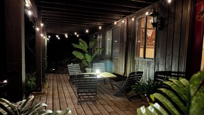 An outdoor patio with wooden flooring, a table, and chairs, surrounded by potted plants and string lights. - Quintessential Berkshire Escape - Artist Home, Tucked Away in Nature (Otis)