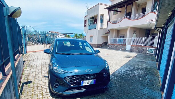 Un'auto blu parcheggiata su un vialetto lastricato, con un muro in pietra da un lato e un edificio con balconi dall'altro.