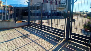 Un cancello chiuso da una recinzione in metallo, terreno pavimentato e un edificio con un balcone sullo sfondo.