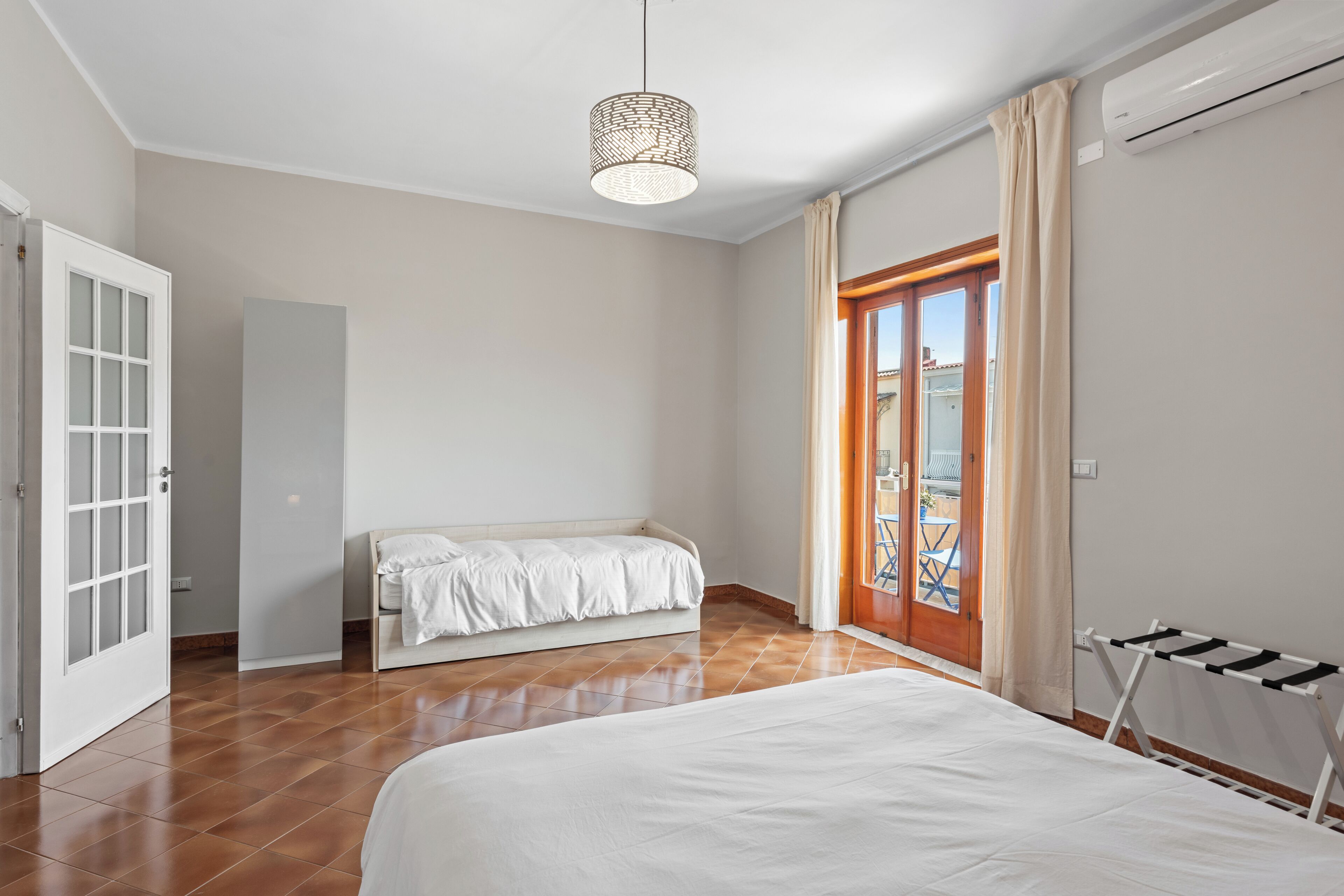 A bedroom with a bed, a bench, a door, and a view of the outside.