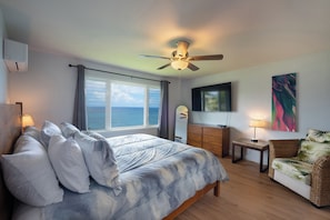 Ein Schlafzimmer mit Bett, Deckenventilator, Fernseher, Kommode, Sessel und Blick auf das Meer.