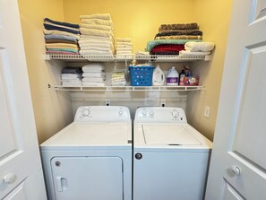 Uma lavanderia com duas máquinas de lavar e secar empilhadas, prateleiras com toalhas dobradas e produtos de lavagem.