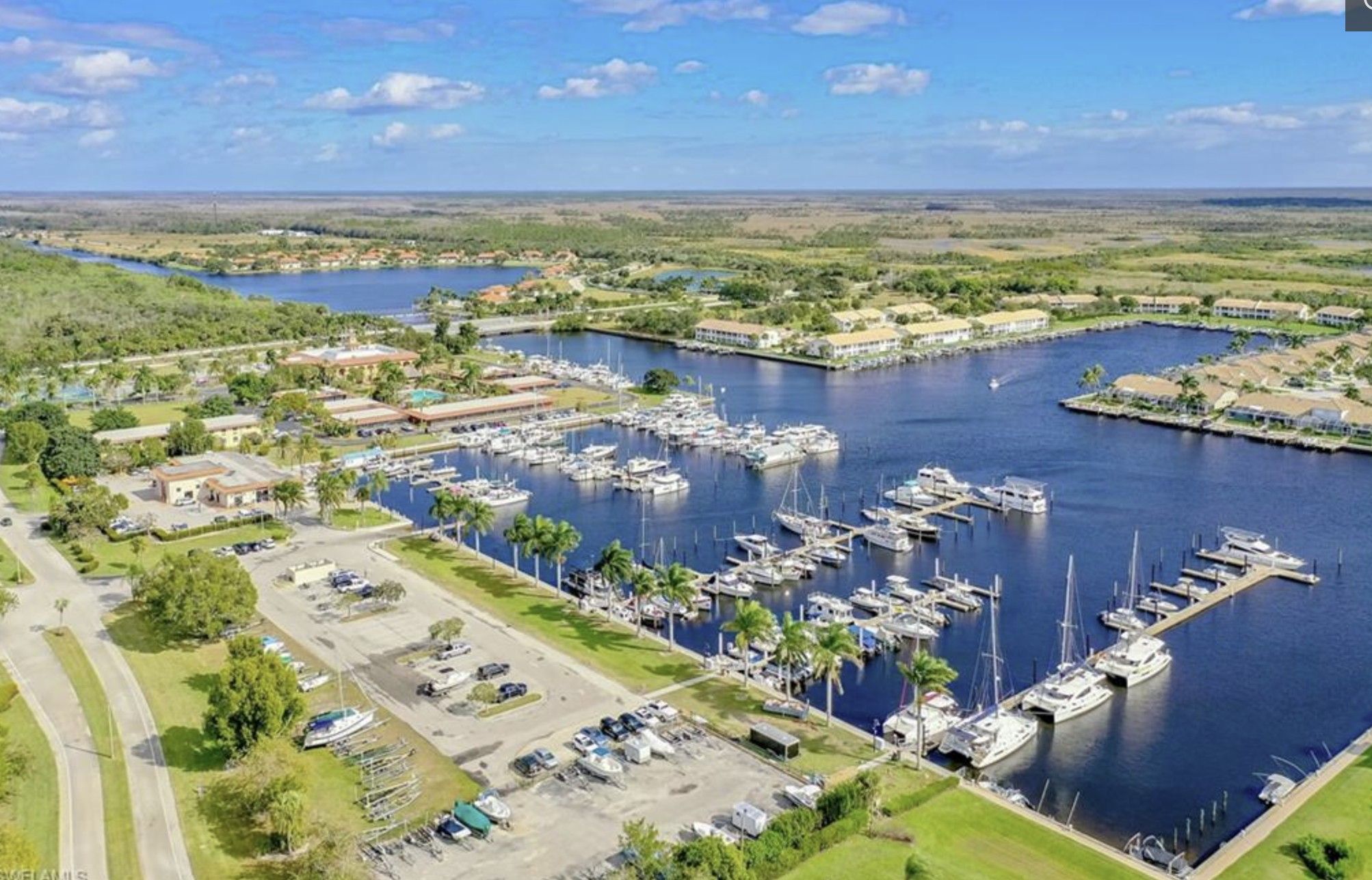 An aerial view of a marina with multiple docks and boats, surrounded by greenery and residential areas.
