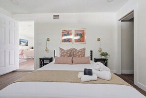 A hotel room with a large bed, two bedside tables, a wall-mounted clock, and a door leading to another room. - Be A Nomad | 3BR With Pool (Jacksonville Beach)