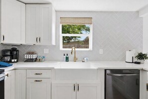 A modern kitchen with white cabinetry, a built-in dishwasher, and a window with a view of trees. - Be A Nomad | 3BR With Pool (Jacksonville Beach)