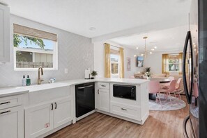 A modern kitchen with white cabinetry, a dishwasher, and a microwave. There is a dining area with a round table and pink chairs. - Be A Nomad | 3BR With Pool (Jacksonville Beach)
