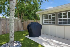 Un jardin avec une clôture en bois, un arbre, un barbecue et une maison avec des fenêtres.