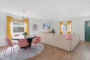 A modern living room with a round dining table, pink chairs, a beige sofa, and yellow curtains. - Be A Nomad | 3BR With Pool (Jacksonville Beach)
