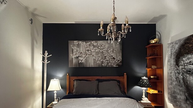 Um quarto com cama, um lustre, mesa de cabeceira, abajur, quadro na parede e uma estante com objetos decorativos.