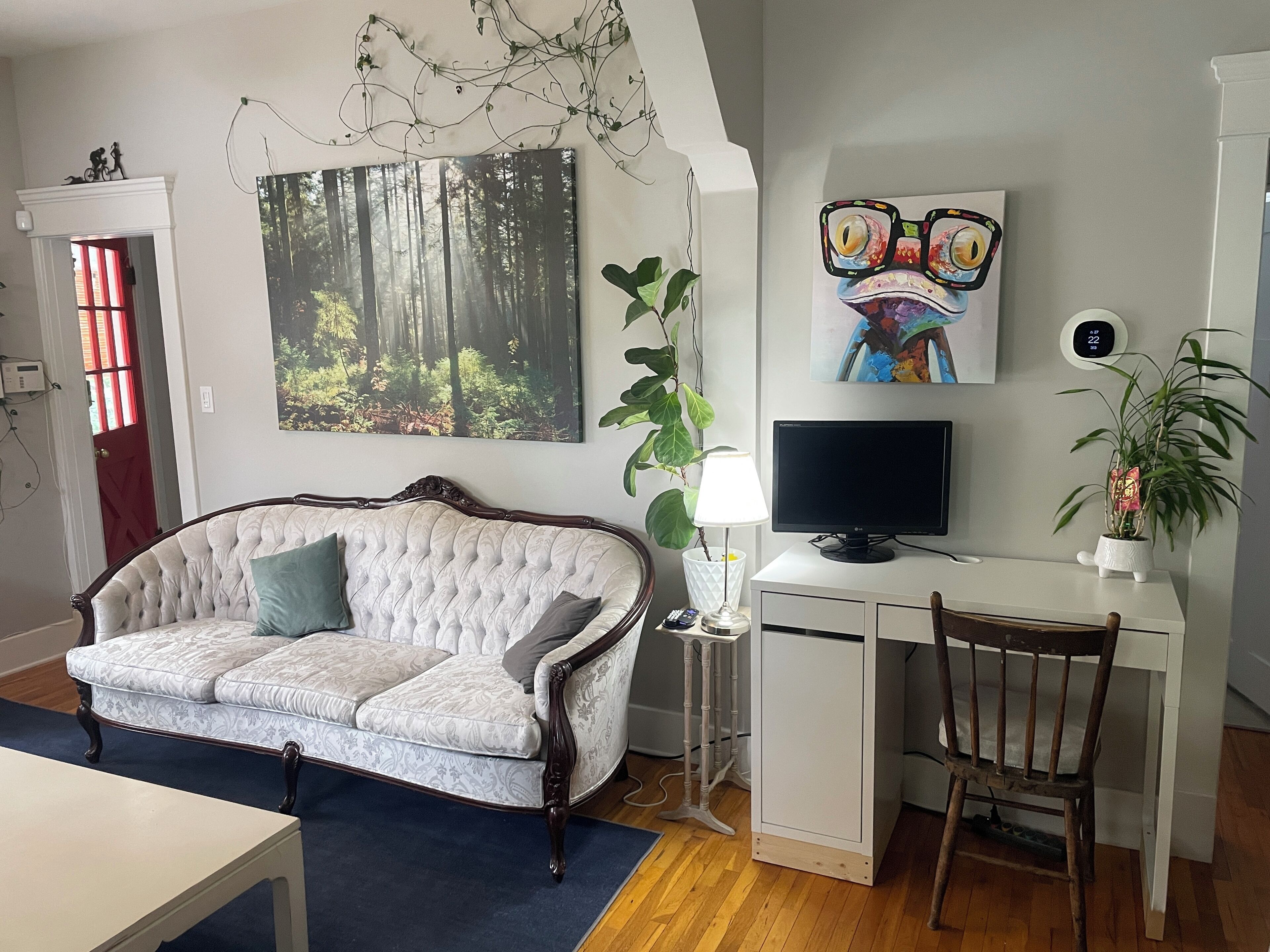 A living room with a tufted sofa, a desk with a computer, a television, and a large forest-themed painting.