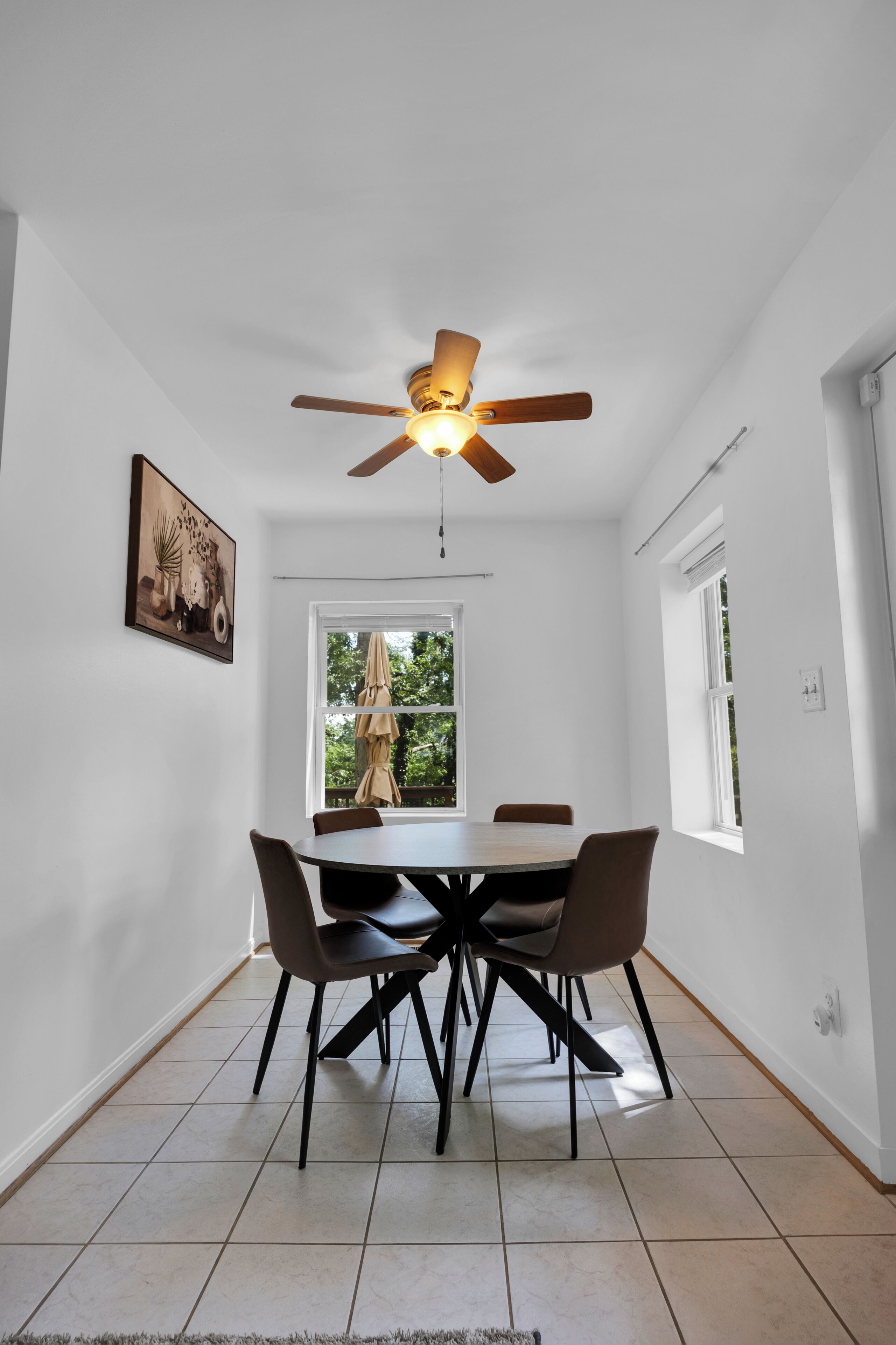 A dining area with a table and chairs, a ceiling fan, a window with a view of trees, and a painting on the wall.