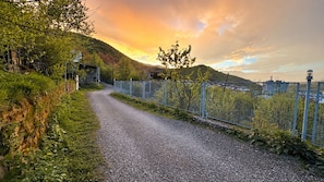 Un chemin de gravier bordé d’une clôture métallique, qui mène à un paysage vallonné illuminé par le coucher du soleil.