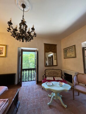 A room with a chandelier, a bed, a table, a chair, a fireplace, and a balcony with a view of greenery.