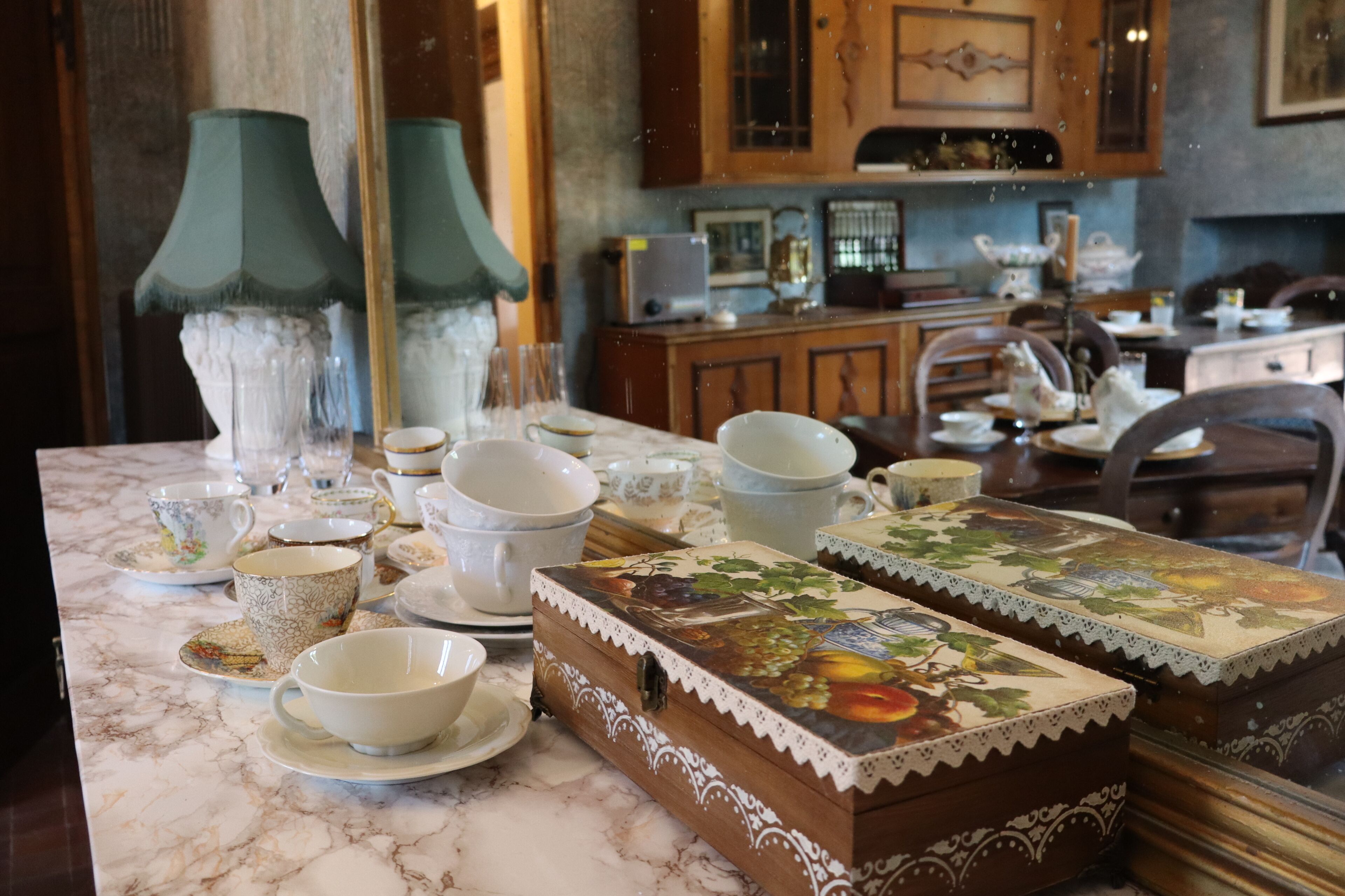 A marble countertop with various tea cups and saucers, a decorative box with a fruit design, and a green lamp.