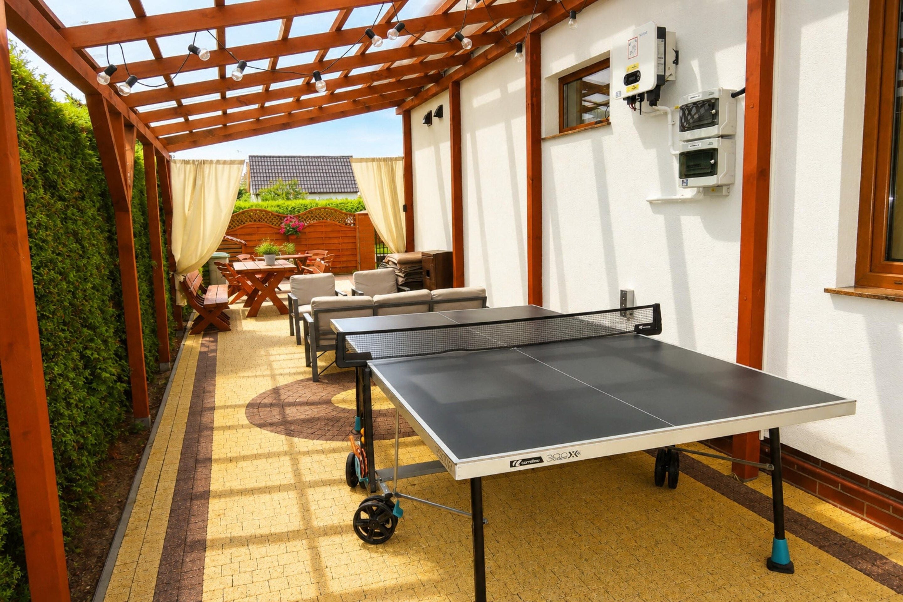 A ping pong table in a covered outdoor area with a pergola, seating, and a view of a house in the background.