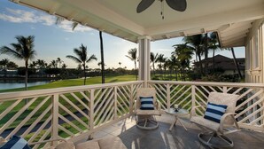 A covered outdoor seating area with two chairs and a table, overlooking a well-maintained lawn and palm trees. - Hilton Pool Pass for 2026!  Lake Views.  M2 Waikoloa Fairway Villas (Waikoloa Beach Resort)