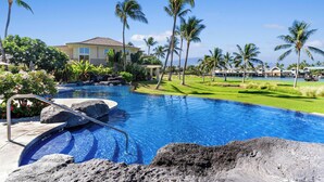 A swimming pool with a clear blue water feature, surrounded by tropical vegetation and a large building in the background. - Hilton Pool Pass for 2026!  Lake Views.  M2 Waikoloa Fairway Villas (Waikoloa Beach Resort)