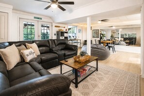 A living room with a sectional sofa, a coffee table, and a bean bag chair. There is a dining area in the background with a table and chairs.