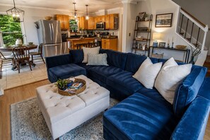 A modern living room with a sectional sofa, a dining area with wooden chairs, and a kitchen with wooden cabinets and stainless steel appliances.