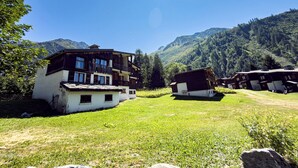 En traditionell byggnad i chaletstil med ett sluttande tak, omgiven av frodig grönska och berg i bakgrunden.