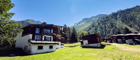 A traditional chalet-style building with a sloped roof, surrounded by lush greenery and mountains in the background.