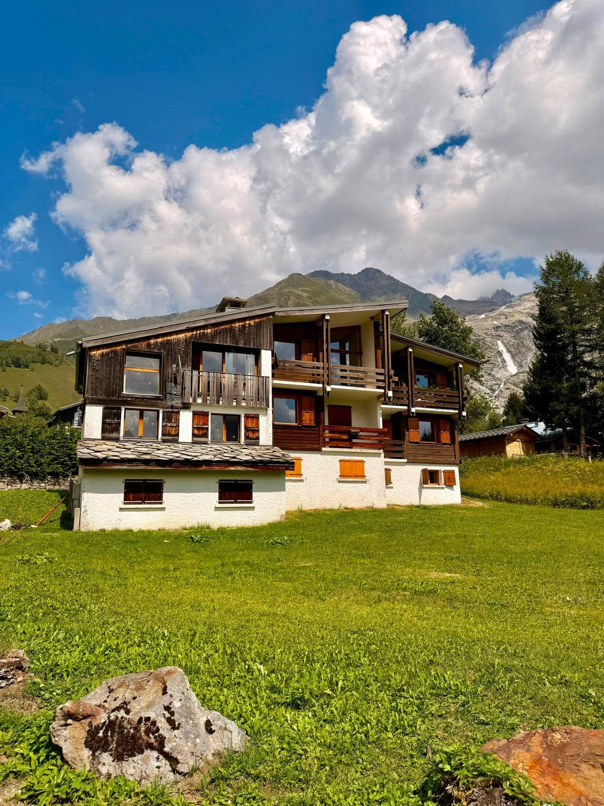 Un chalet de dos pisos con balcón, ubicado en un campo cubierto de hierba y montañas al fondo.