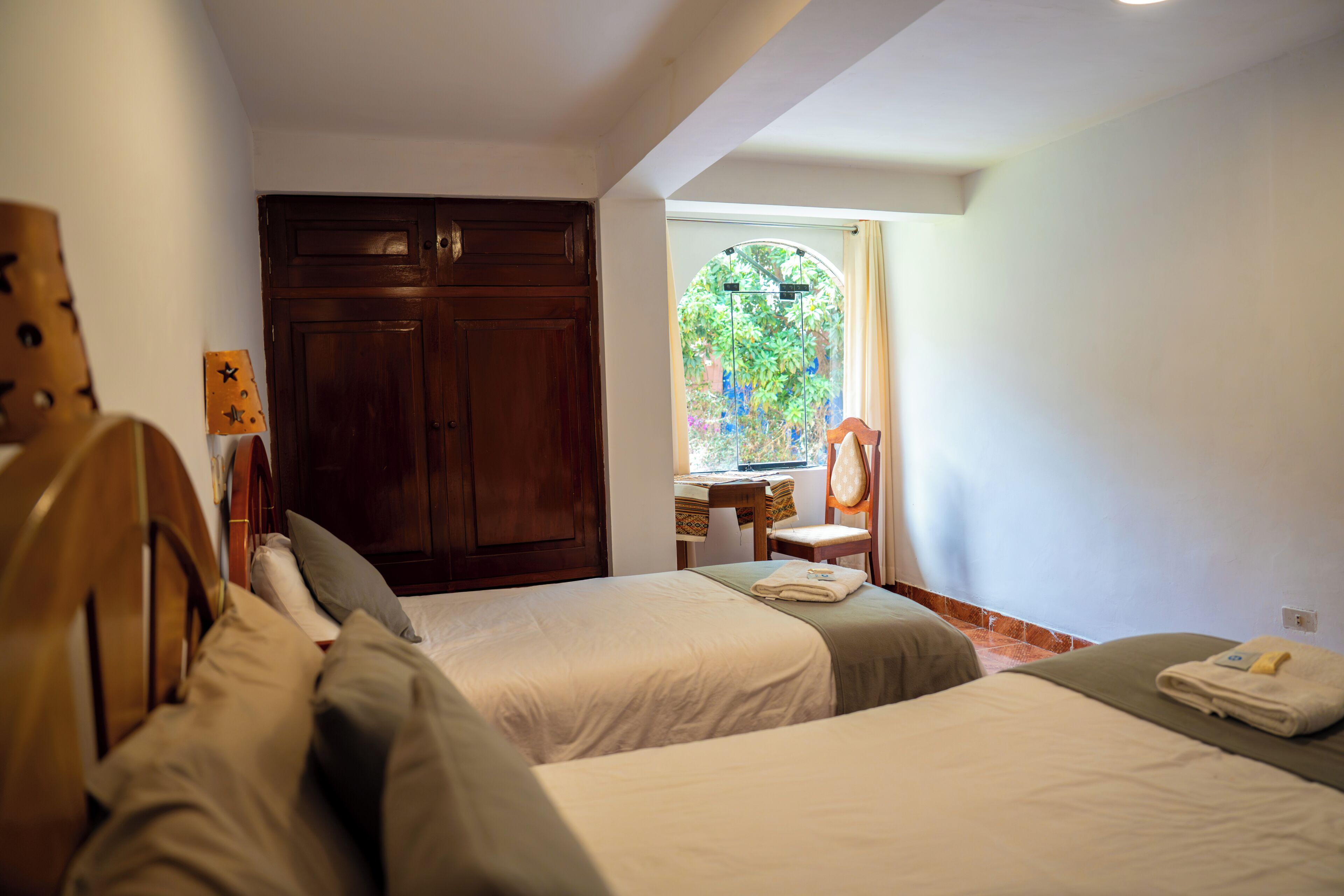 Une chambre avec deux lits, une armoire en bois, une petite table avec une chaise et une fenêtre donnant sur un paysage verdoyant.