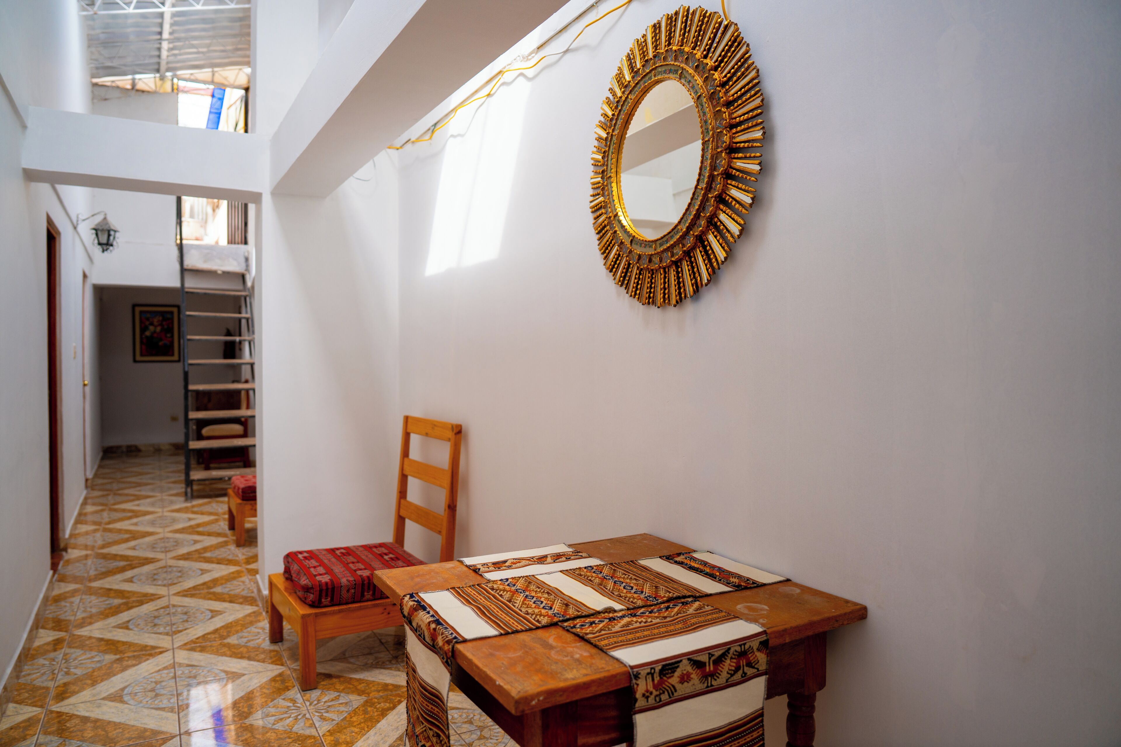 Una habitación con una mesa de madera, un banco estampado, un espejo y una escalera que sube a un nivel superior.