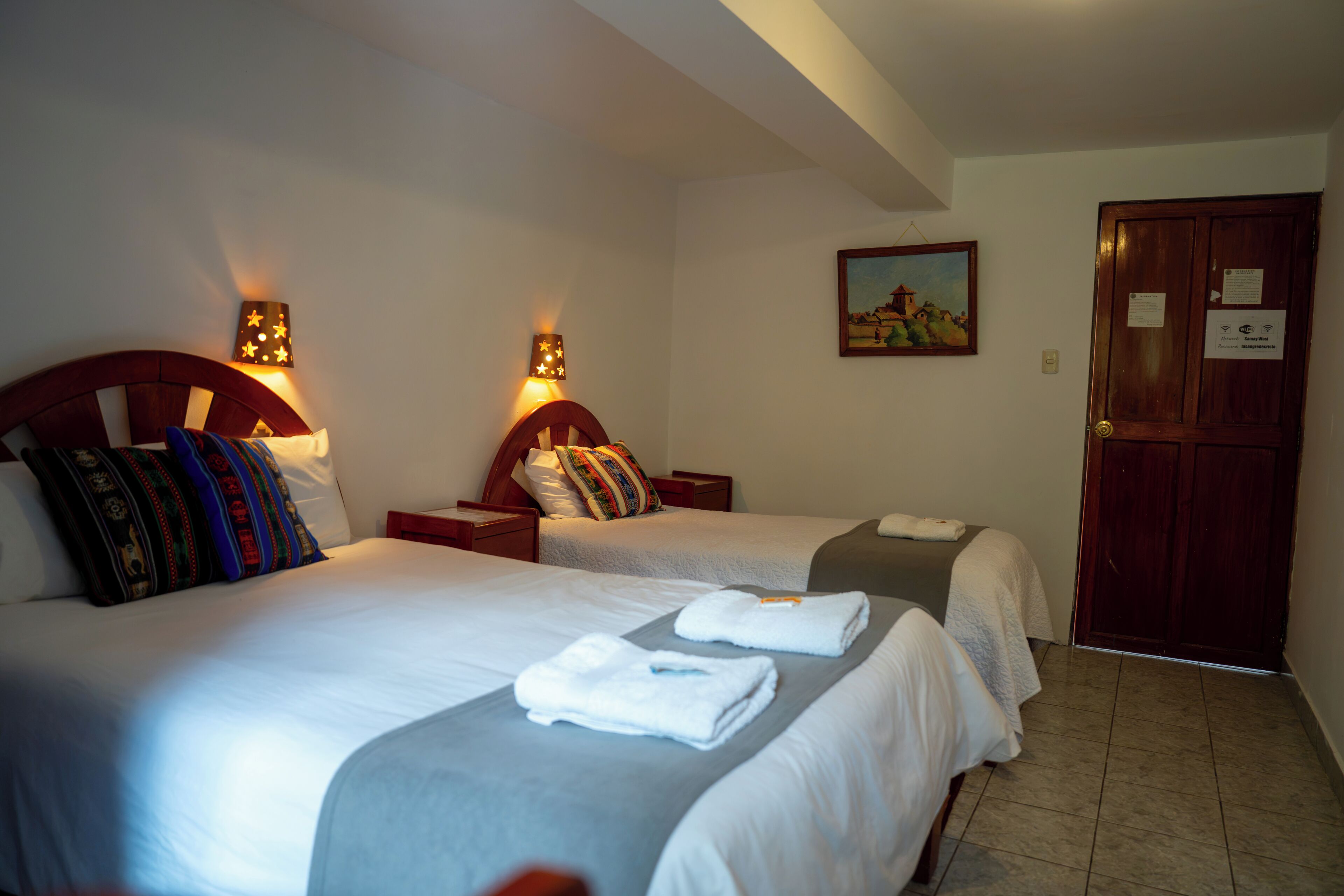 Une chambre d’hôtel avec deux lits, chacun doté d’une tête de lit et de lampes de chevet. On y trouve une porte avec une pancarte, un tableau au mur et un sol carrelé.