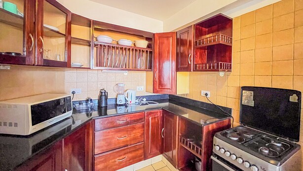 A kitchen with wooden cabinets, a microwave, a stove, and a countertop with various appliances. - Mvuto mzuri Beachfront Apartments (Mombasa)