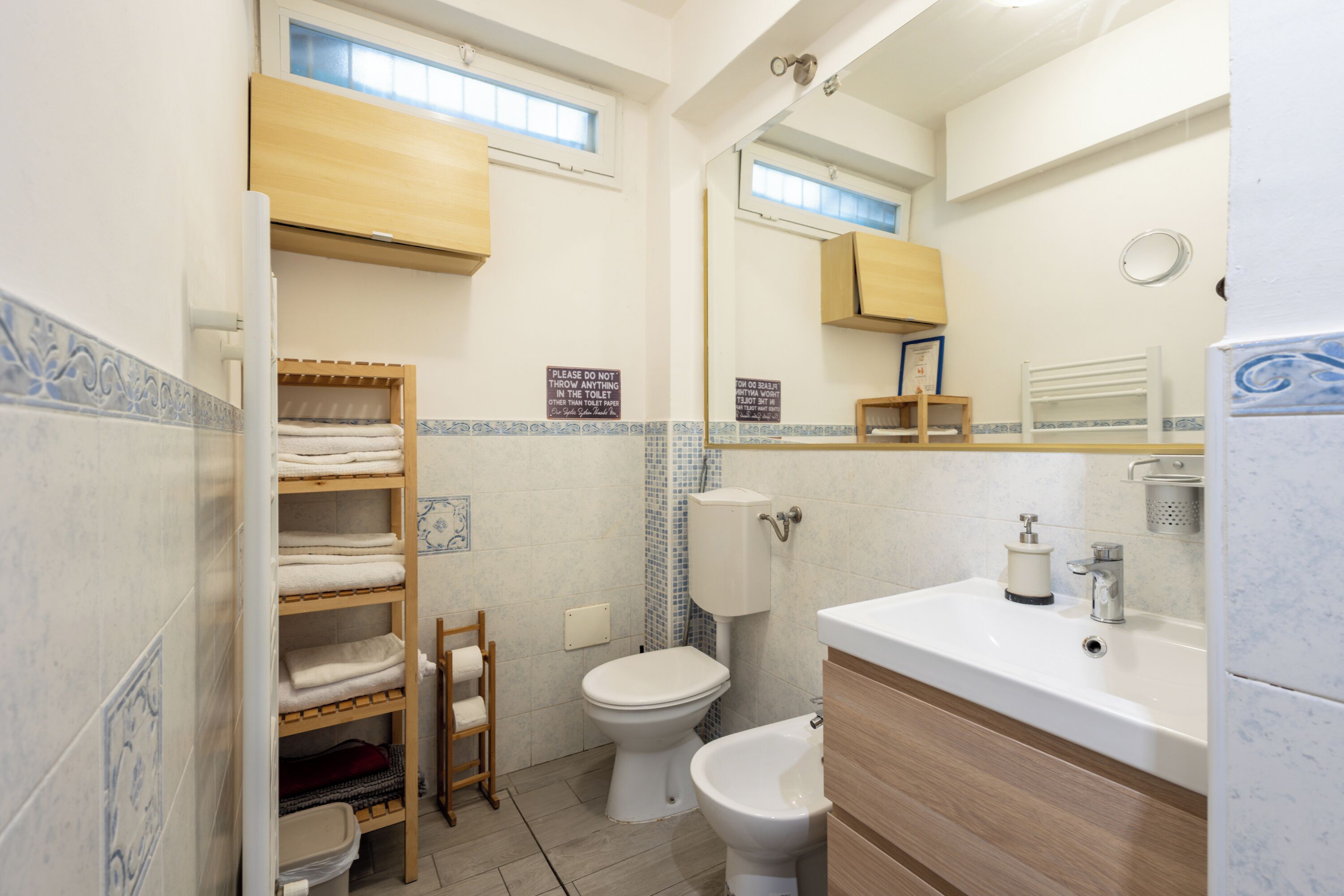 Ein Badezimmer mit Toilette, Bidet und Waschbecken mit Spiegel darüber. Handtücher liegen auf einem Regal und ein Holzschrank befindet sich über dem Waschbecken.