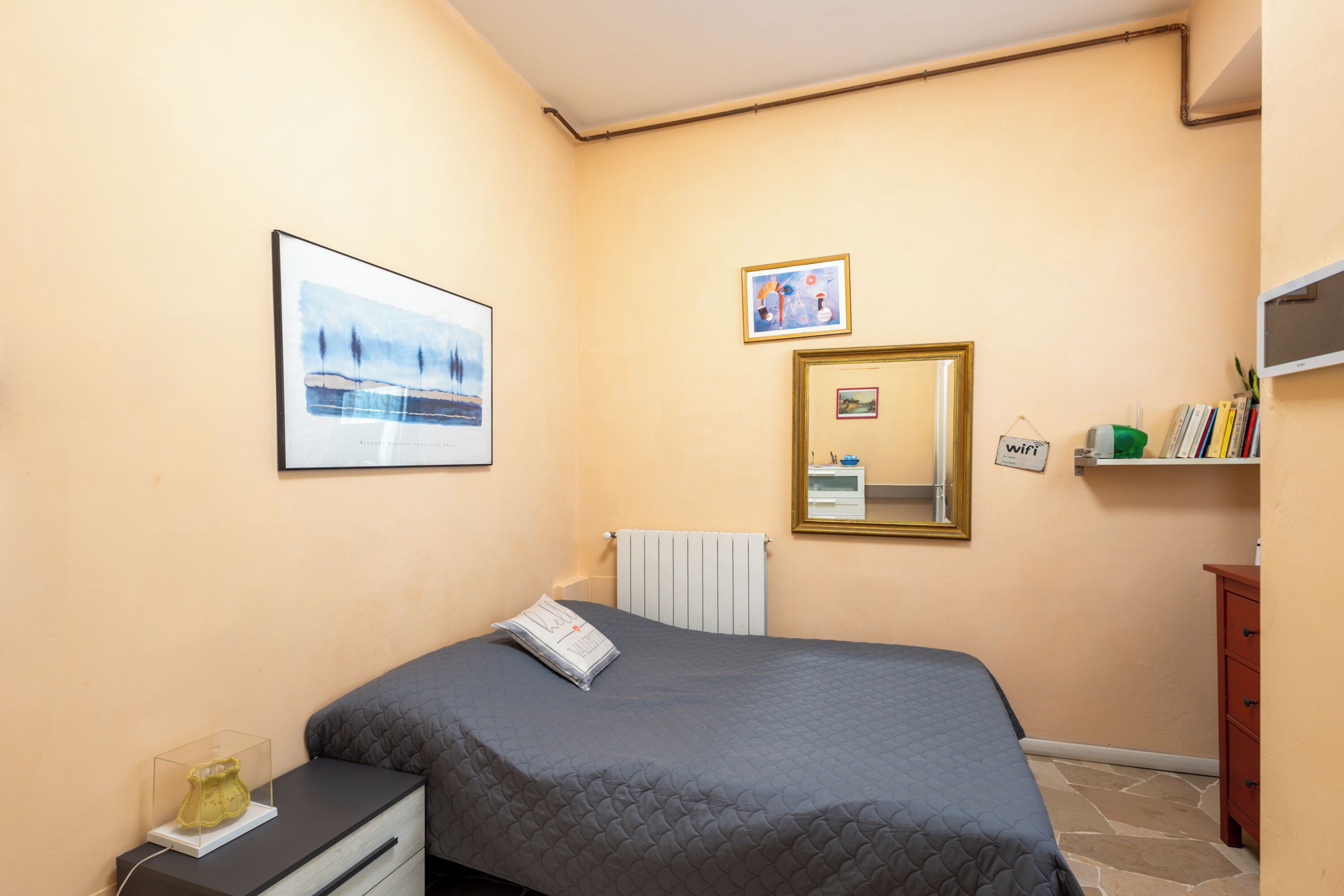 Une chambre à coucher avec un lit, une table de chevet, un miroir et une étagère murale contenant des livres.