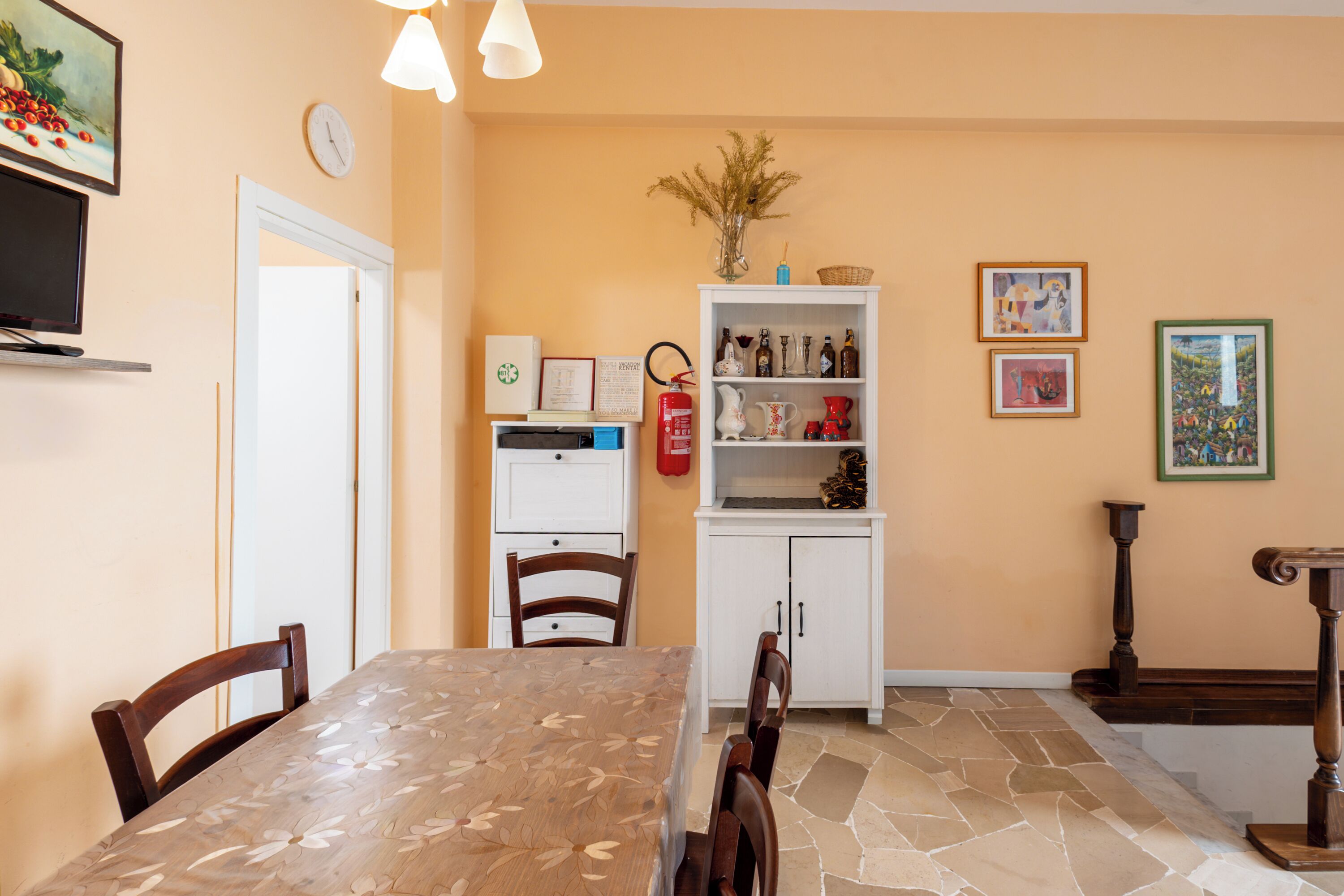 Un comedor con una mesa y sillas, un armario blanco con objetos decorativos, un extintor y una obra de arte en la pared.
