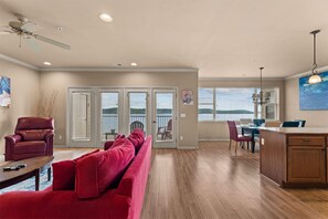 A spacious living area with a red sofa, a leather chair, and a dining table with chairs. There is a kitchen area with wooden cabinets and a pendant light. - The Red Curtain Lakefront (Hollister)