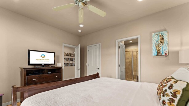 Una camera d'albergo con un letto, una televisione che mostra 'GRAND WELCOME', un comò in legno, una ventola a soffitto e una porta che dà su un bagno.