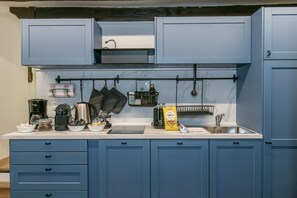 Eine Küche mit blauen Schränken, einer Arbeitsplatte mit verschiedenen Geräten und Kochutensilien sowie einem Hängegestell für Töpfe und Pfannen.