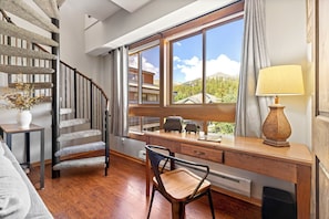 A modern room with a wooden desk, a chair, a staircase with a unique design, and a large window offering a view of buildings and trees. - Walk to Main St & BreckConnect Gondola • Hot Tub & Views • Comfortably Sleeps 10 (Breckenridge)