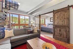 A spacious living area with a large sofa, a coffee table, and a view of a forest through large windows. - Walk to Main St & BreckConnect Gondola • Hot Tub & Views • Comfortably Sleeps 10 (Breckenridge)