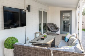 Ein überdachter Außenbereich mit Sitzgelegenheit, einem an der Wand montierten Fernseher, einem gepolsterten Sessel, einer Couch und einem kleinen Tisch mit einer Pflanze.