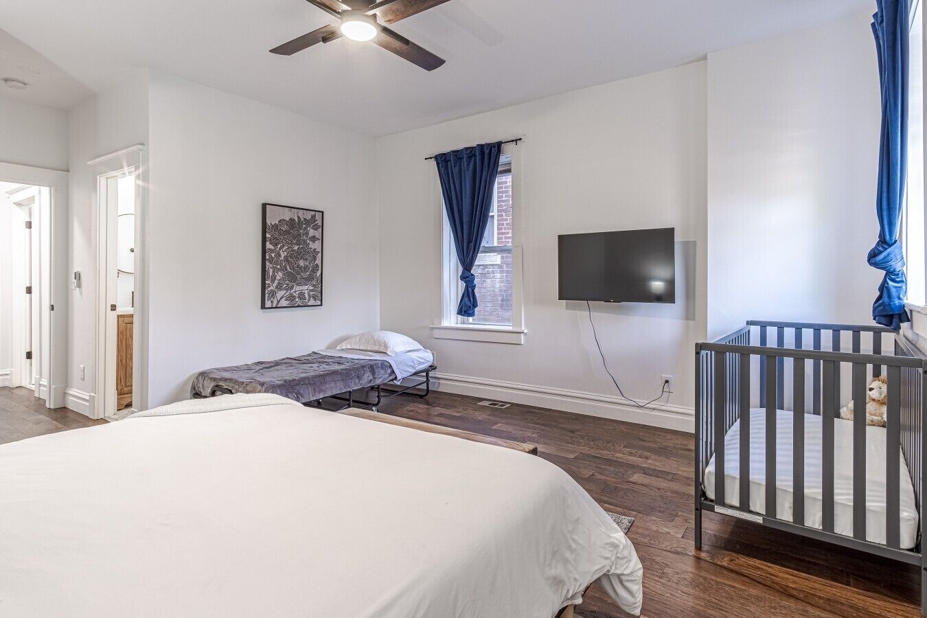 A bedroom with a large bed, a smaller bed, a crib, a ceiling fan, a TV, and a window with curtains.
