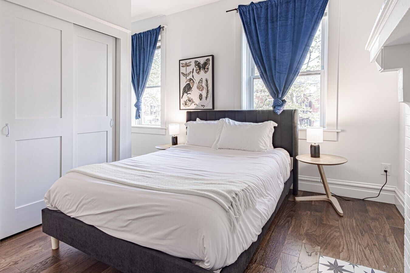 Een slaapkamer met een bed, twee nachtkastjes, een raam met blauwe gordijnen en een kunstwerk aan de muur.