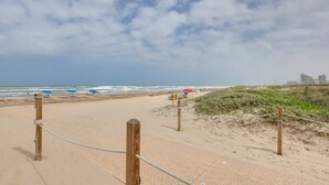 Ein Strand mit einem sandigen Pfad, der zum Wasser führt, einigen Sonnenschirmen und einer entfernten Stadtlandschaft.